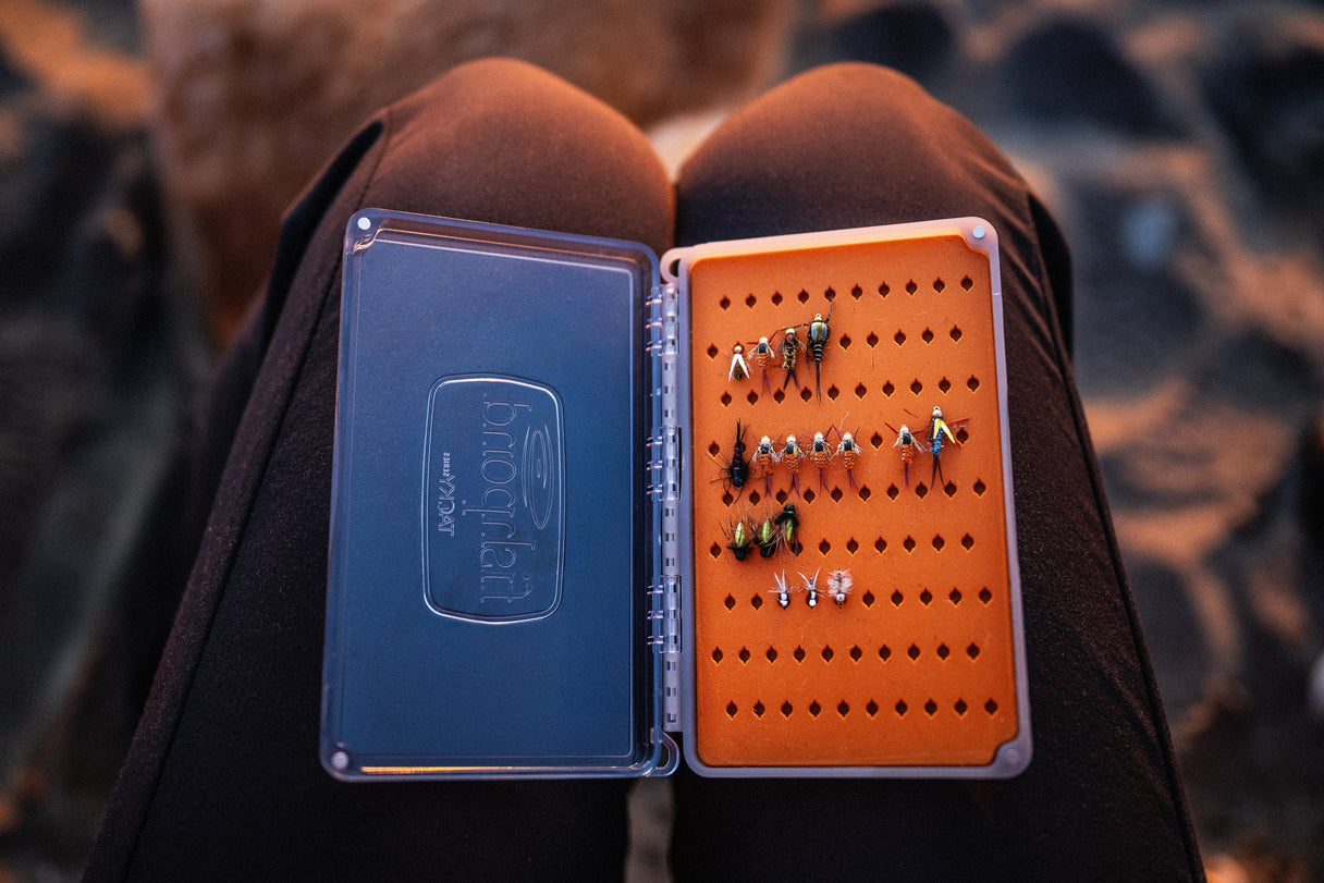 Top-down view of Fishpond Tacky Pescador medium fly box with clear lid and orange dynamic foam holding assorted flies on an angler's knees