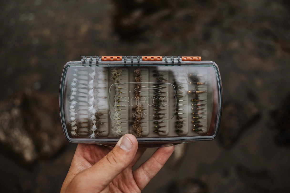 Top view of Fishpond Tacky Pescador medium fly box with clear lid, orange latches and tacky foam filled with flies, held over rocks