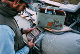 Angler holding open Tacky Pescador medium clear fly box showing tacky foam and flies beside a Fishpond bag on a frosty boat