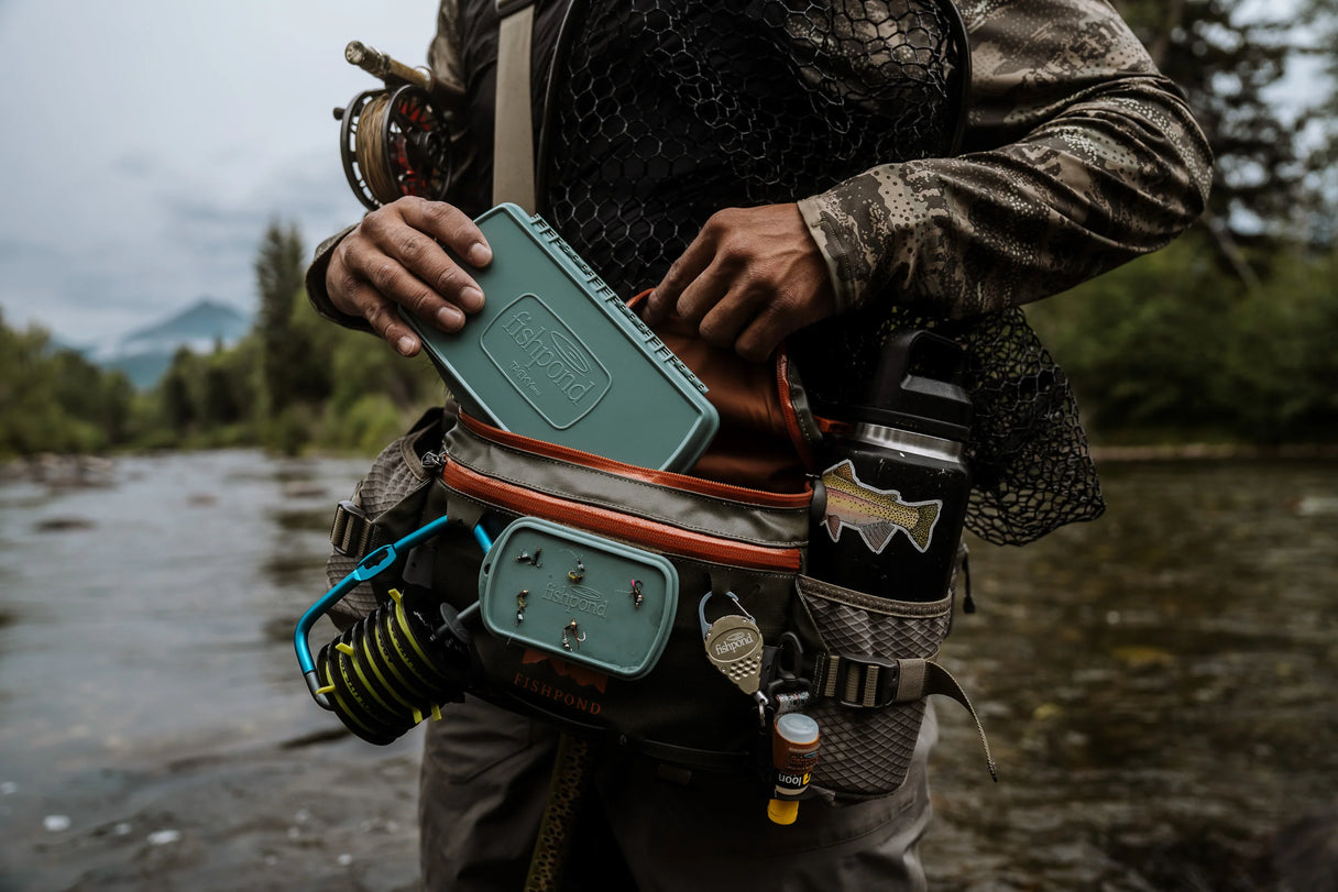 Angler placing a Fishpond Pescador large smoke grey fly box into a waist pack while standing in a river