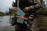 Angler placing a Fishpond Pescador large smoke grey fly box into a waist pack while standing in a river