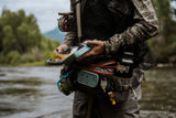 Angler holding open smoke grey Fishpond Tacky Pescador large fly box showing assorted flies while fly fishing at a river
