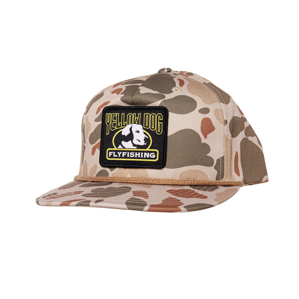 A flat-brim snapback cap in a desert camouflage pattern, featuring a black rectangular patch on the front with the text 'YELLOW DOG FLYFISHING' and a dog graphic.
